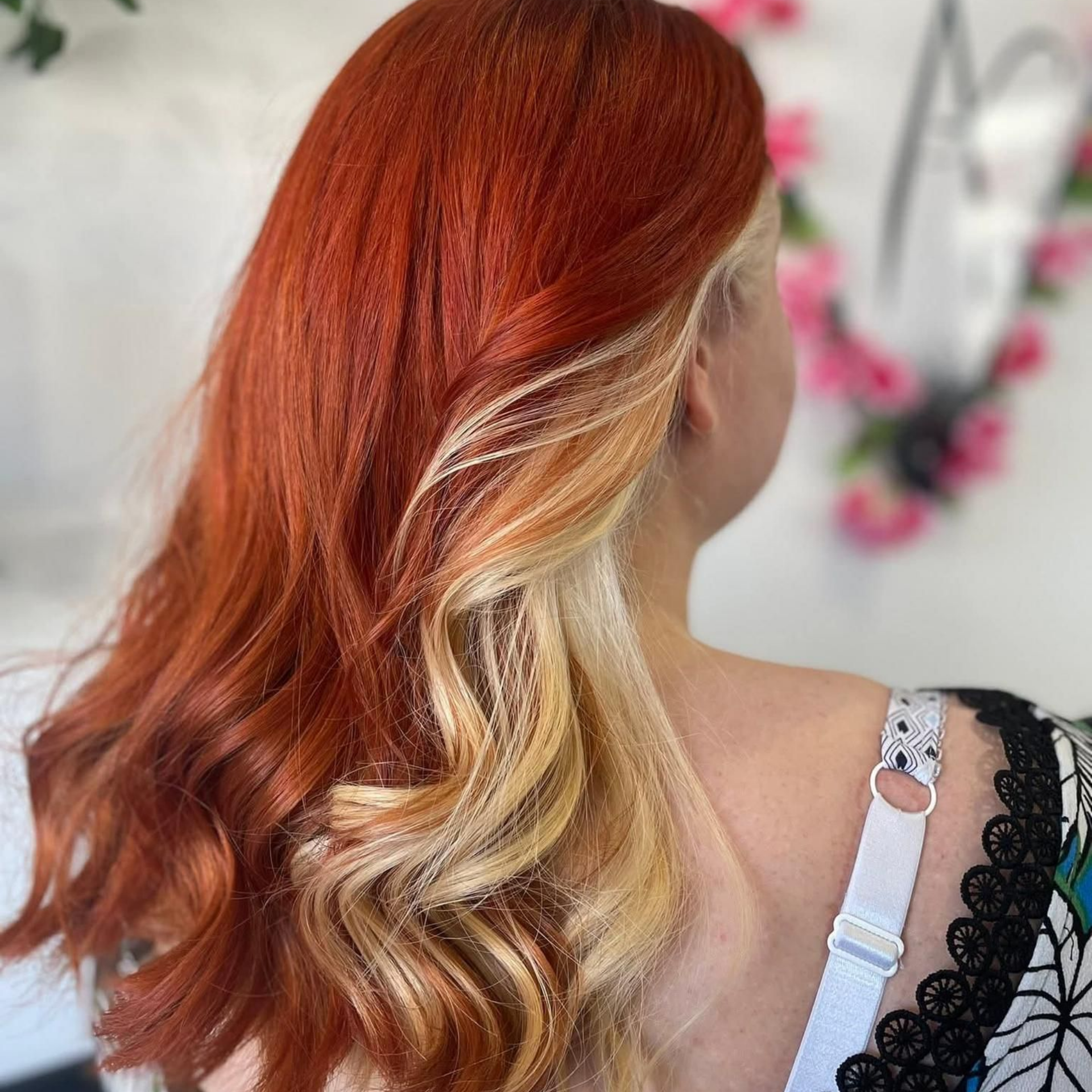 Cheveux ondulés d'une femme : roux flamboyant sur le dessus, blond aux pointes. Bretelle et décor floral en arrière-plan.