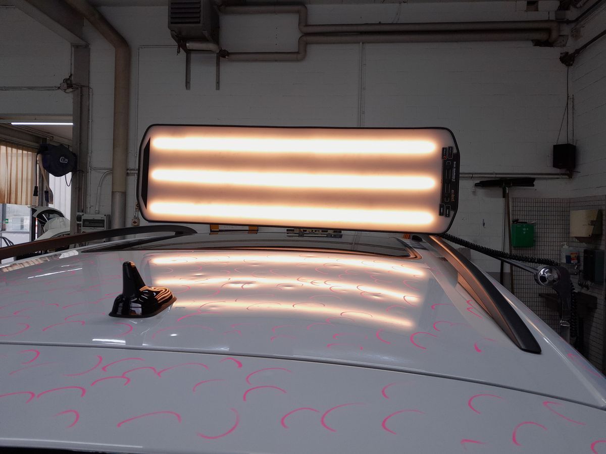 Ein Mechaniker inspiziert in einer Garage den Scheinwerfer eines silbernen Autos und hält dabei ein Klemmbrett in der Hand.