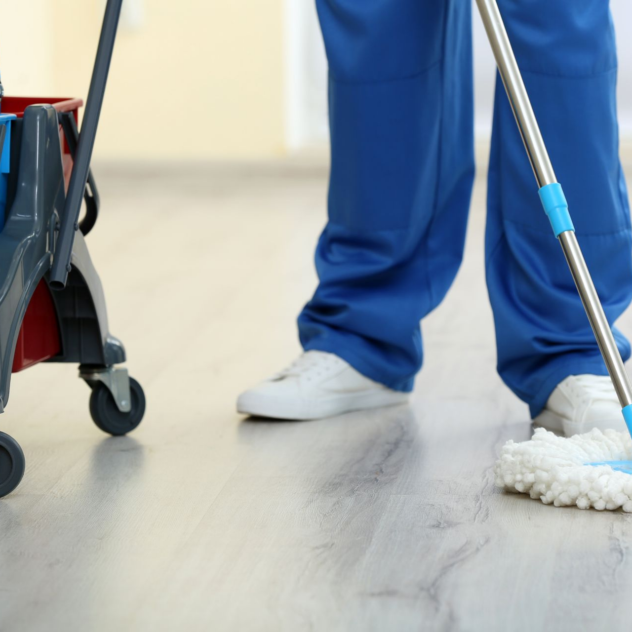 Personne en bleu nettoyant le sol avec une serpillière ; chariot de nettoyage à gauche.