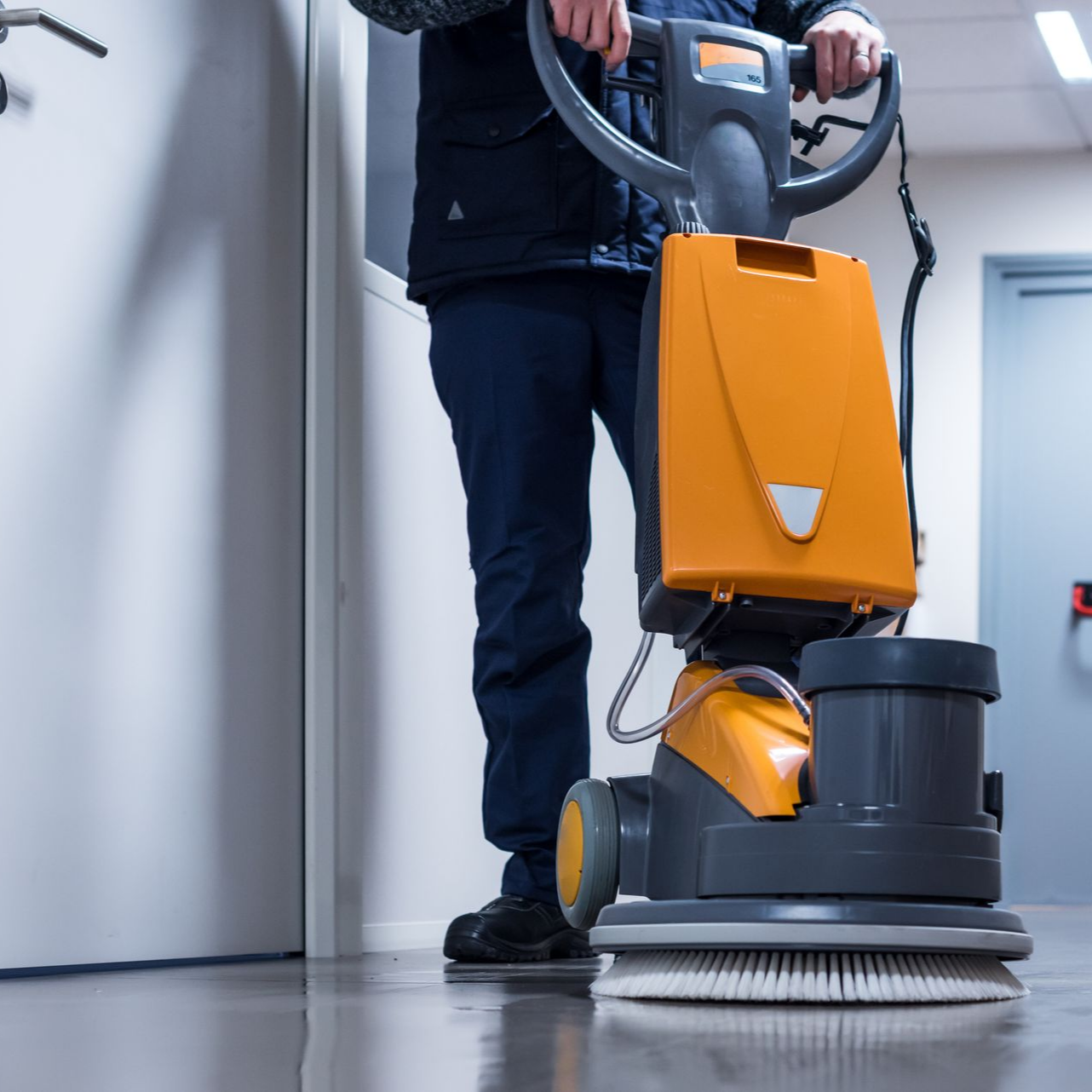 Une personne utilise une machine de nettoyage de sol orange dans un couloir.