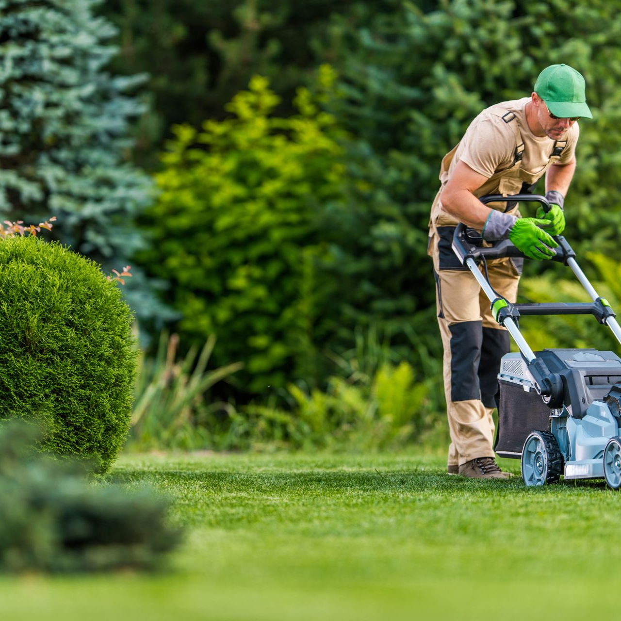 Un homme tond une pelouse verte dans un jardin, portant une casquette verte, une salopette beige et des gants.