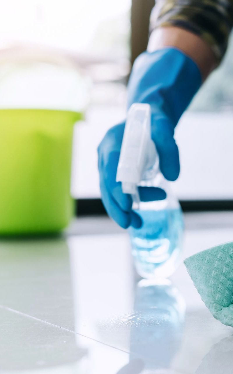 Une personne portant des gants bleus vaporise un produit nettoyant sur une surface carrelée blanche, à côté d'un seau vert et d'une éponge.