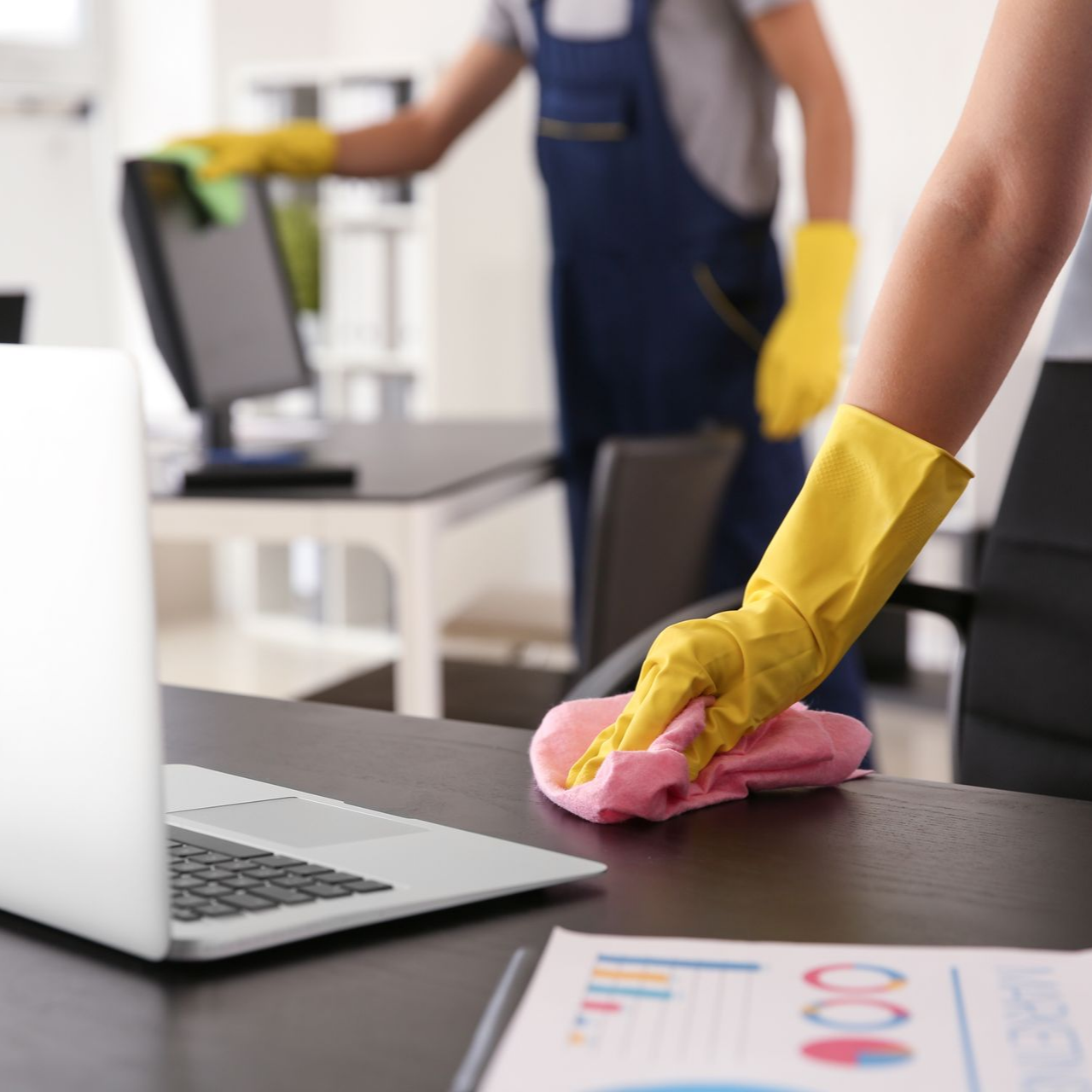 Une personne portant des gants jaunes essuie un bureau, une autre nettoie un ordinateur dans un environnement de bureau.