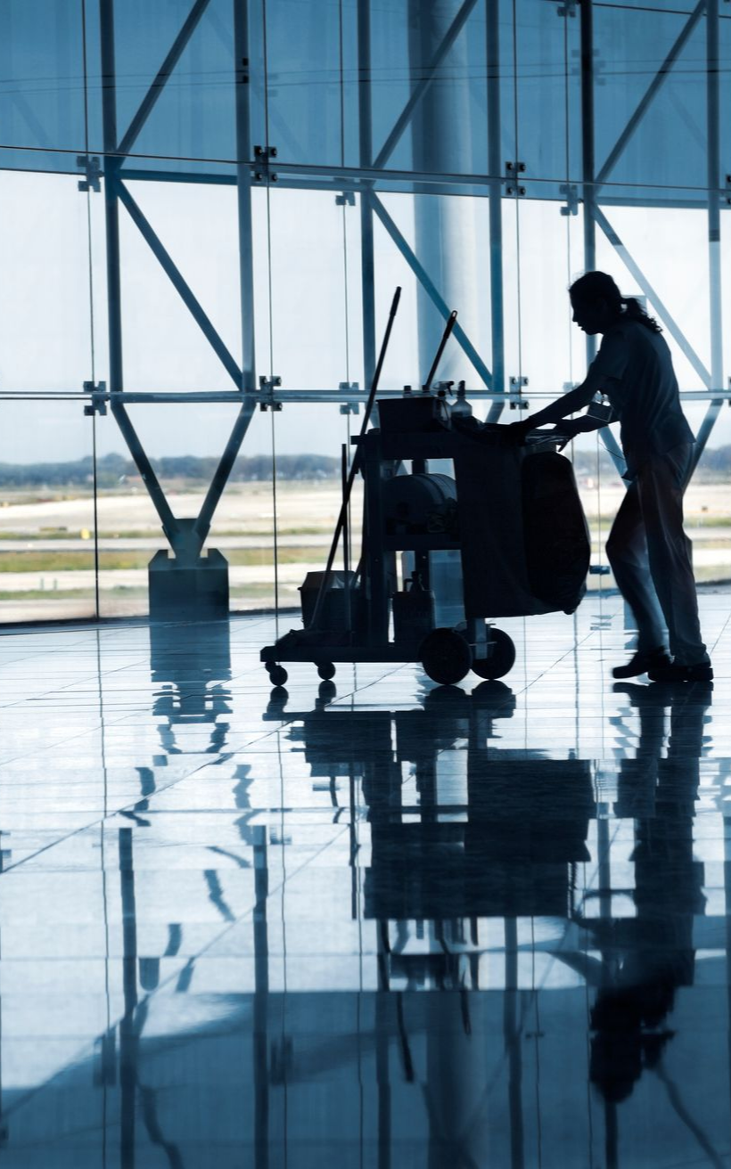 Une personne pousse un chariot de nettoyage sur un sol brillant dans un grand bâtiment moderne doté de grandes fenêtres.