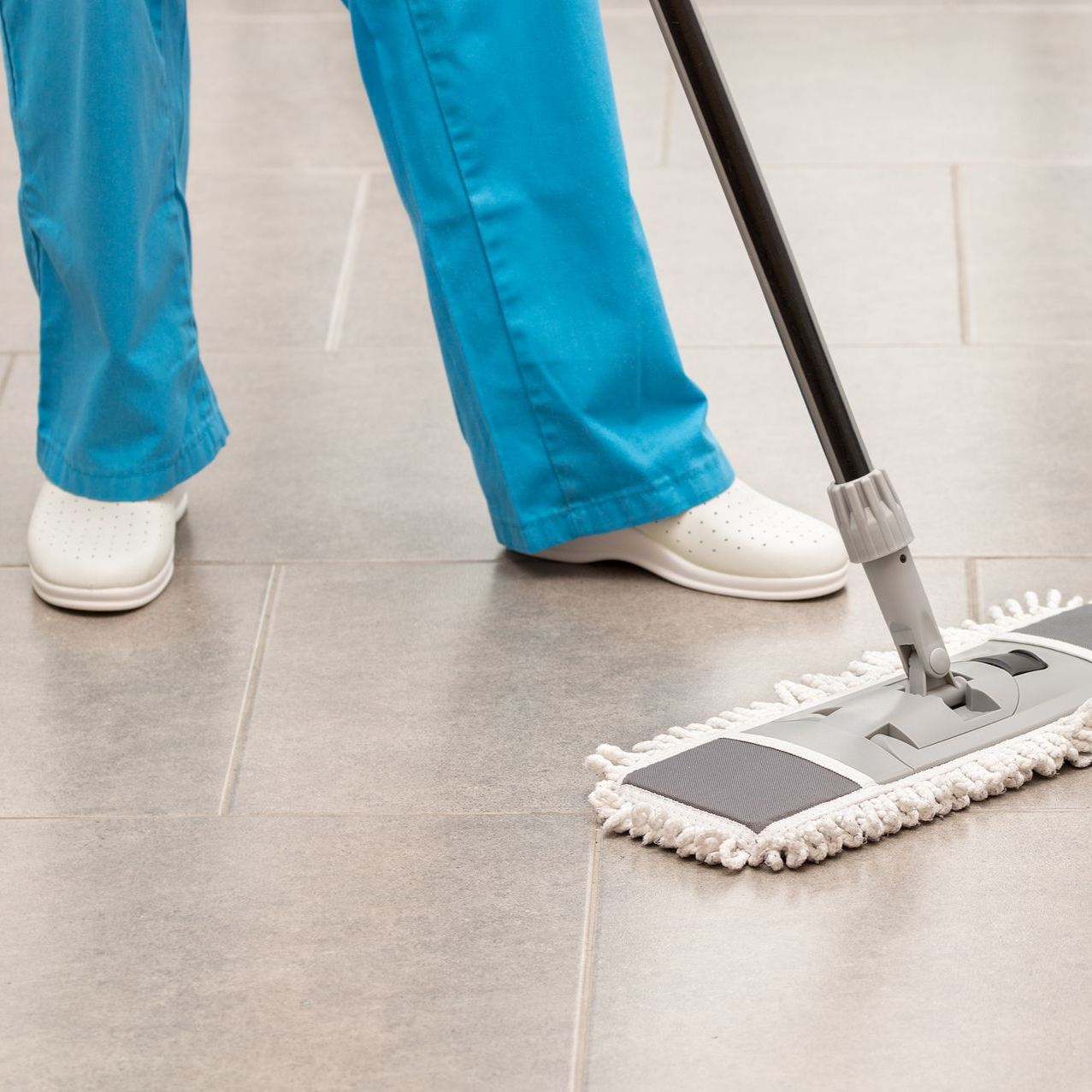 Une personne en blouse bleue lave un sol carrelé avec une serpillière grise.