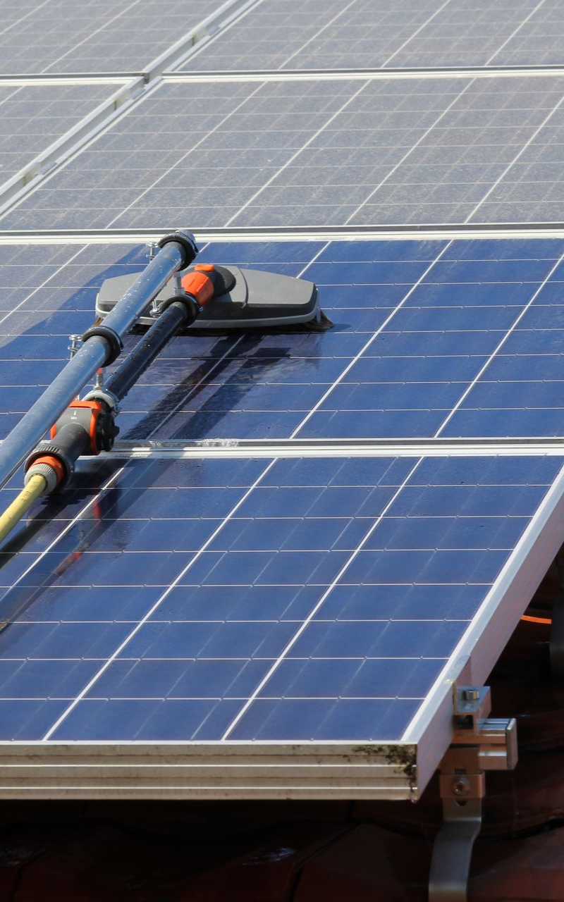 Nettoyage des panneaux solaires avec une brosse à long manche ; les panneaux sont bleus, le toit est marron, journée ensoleillée.