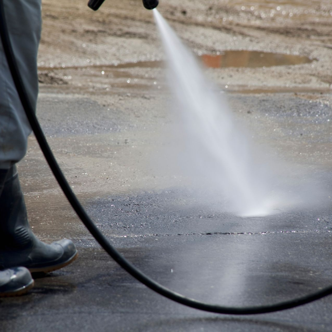 Une personne bottée nettoie une surface en béton au jet d'eau à haute pression, créant ainsi un jet d'eau.