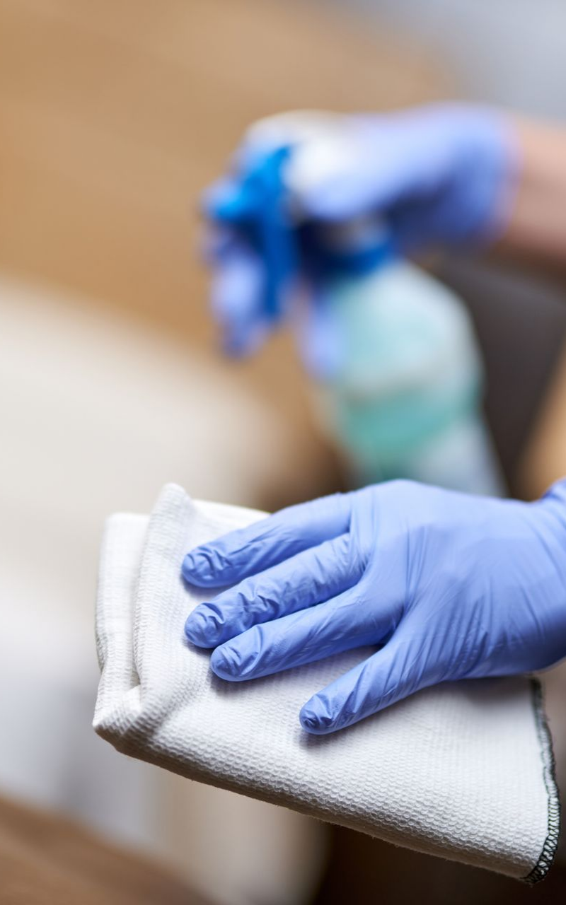 Une personne portant des gants bleus vaporise du produit nettoyant sur un chiffon blanc pour essuyer une surface.
