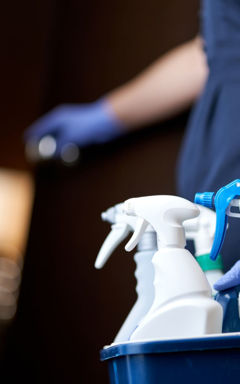 Des produits de nettoyage dans un seau bleu, une personne portant des gants ouvrant une porte sombre.