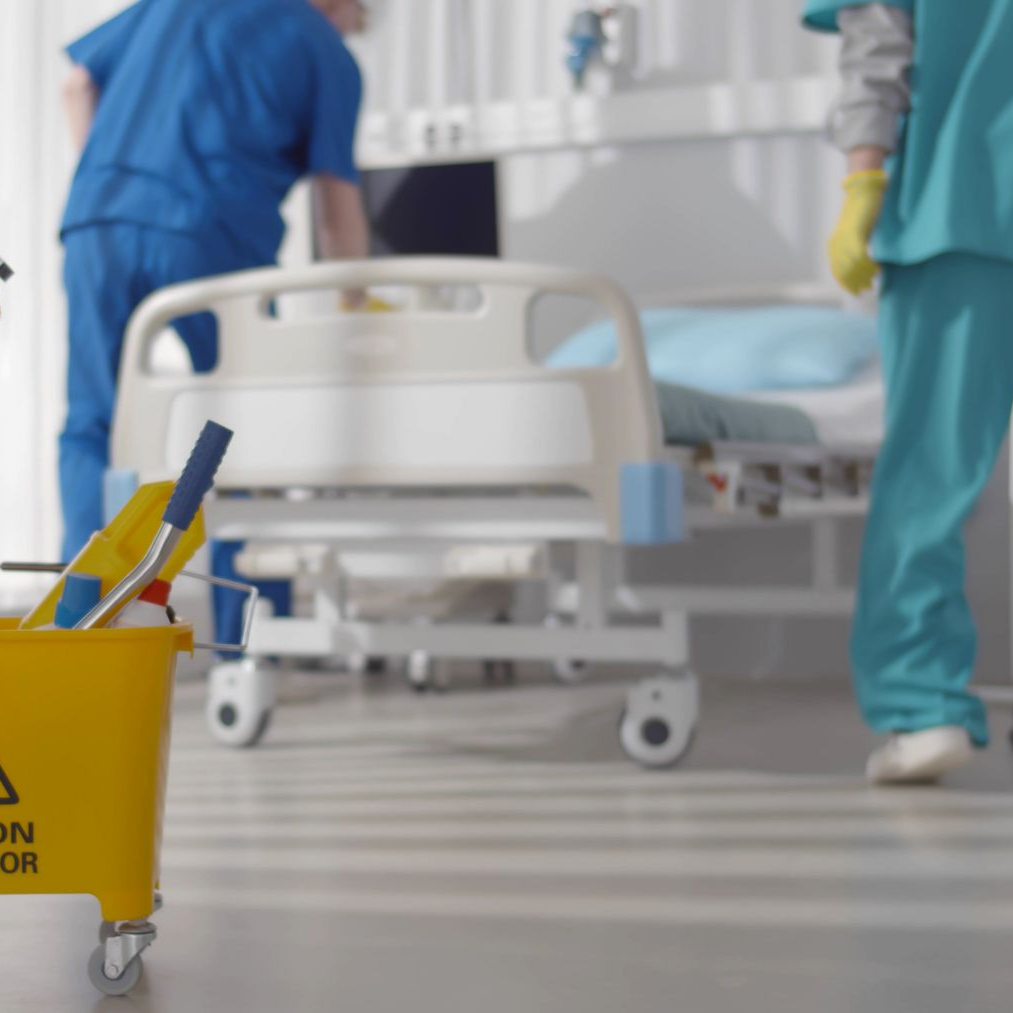 Matériel de nettoyage devant un lit d'hôpital, avec deux soignants en arrière-plan.