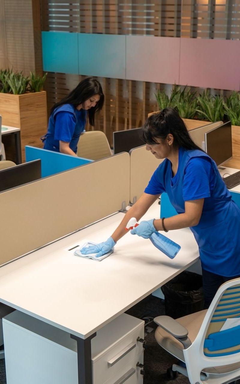 Deux personnes nettoient les bureaux, l'une vaporise et essuie, vêtues d'uniformes bleus et de gants.