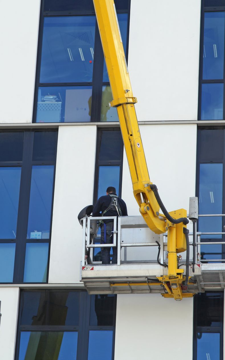Un ouvrier, posté sur une plateforme élévatrice, travaille sur la façade d'un bâtiment blanc moderne aux encadrements de fenêtres bleus.