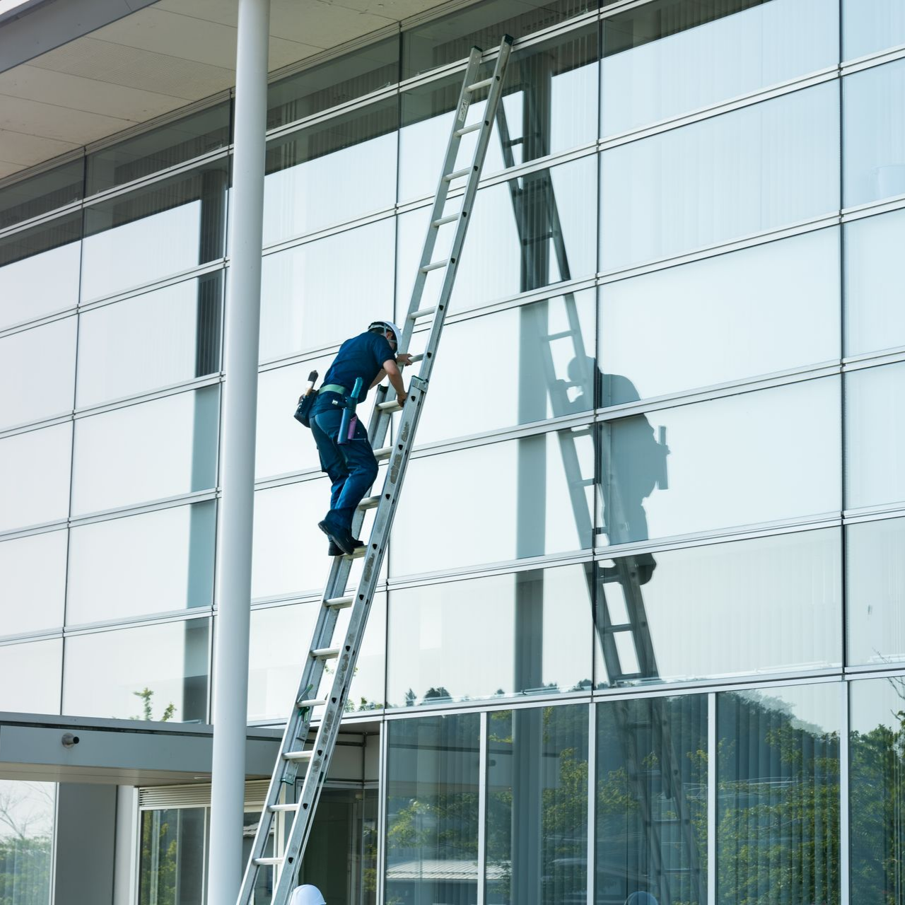 Une personne monte à une échelle pour laver les grandes fenêtres d'un immeuble moderne ; une poutre de soutien blanche est visible.