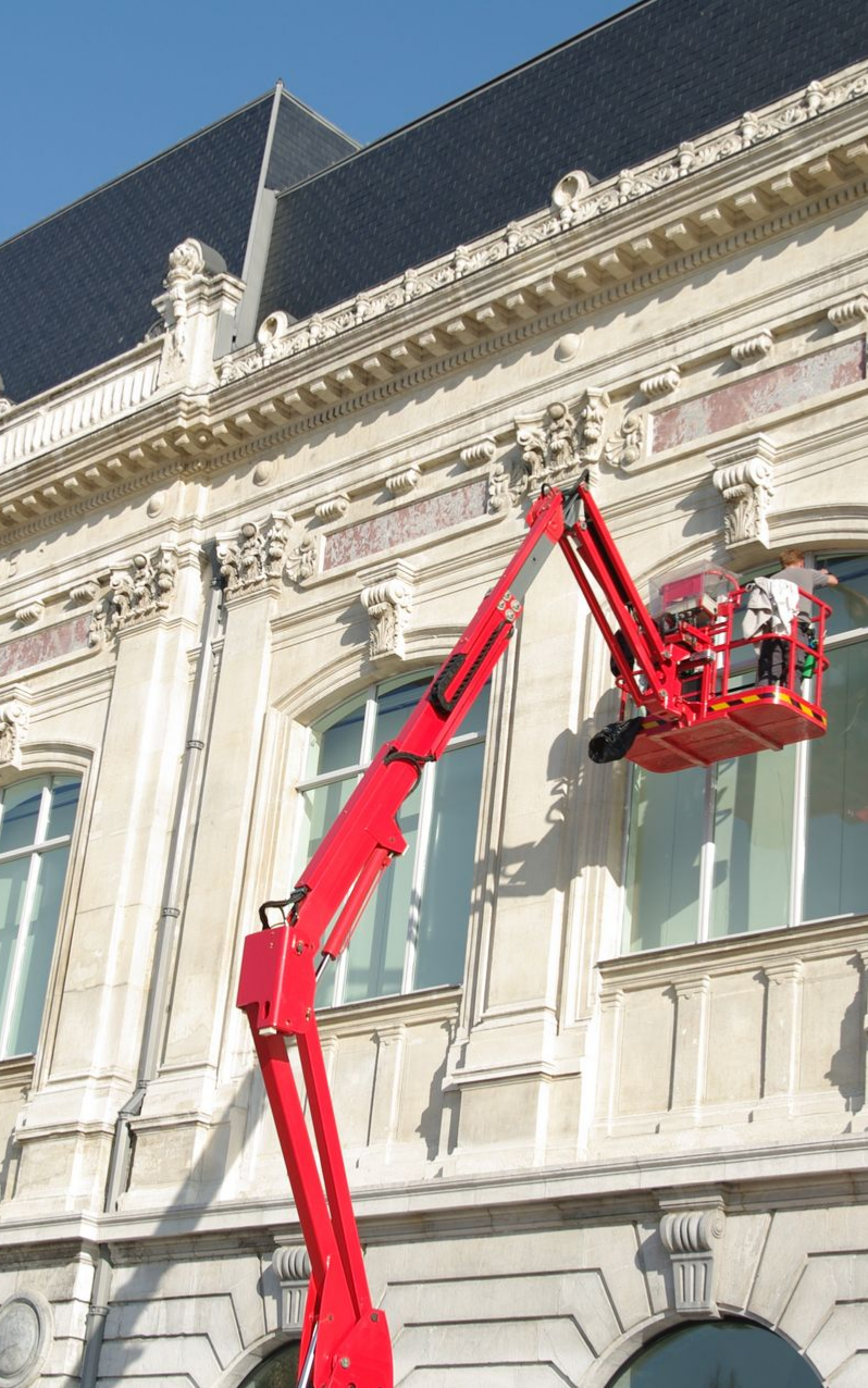 Plateforme élévatrice rouge s'étendant jusqu'à la façade d'un immeuble, un ouvrier sur la plateforme.