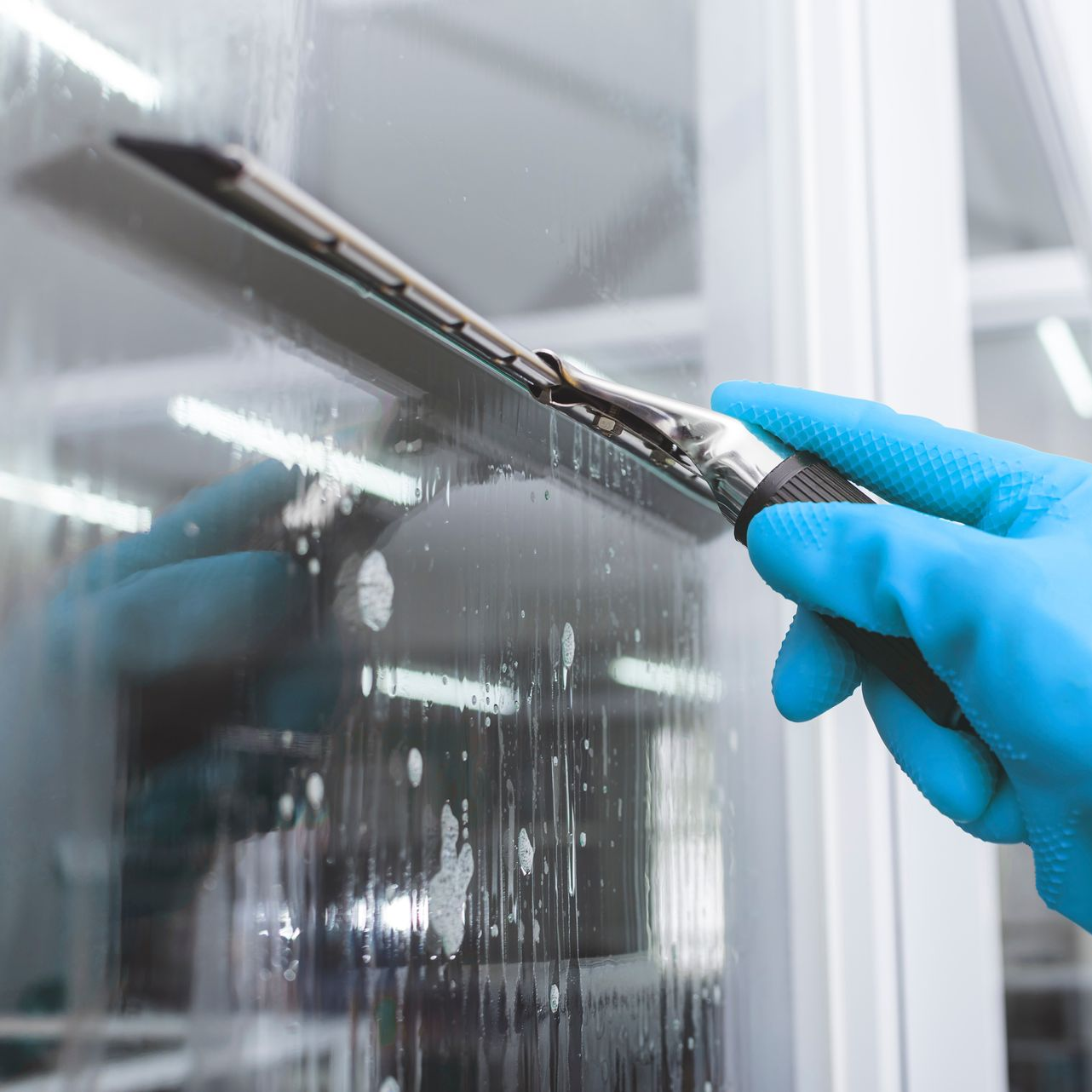 Main gantée de bleu utilisant une raclette pour nettoyer une vitre.