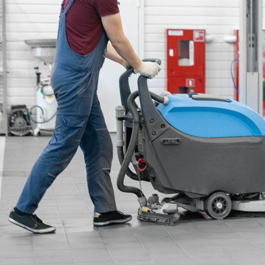 Une personne en salopette bleue utilise une autolaveuse dans un garage.