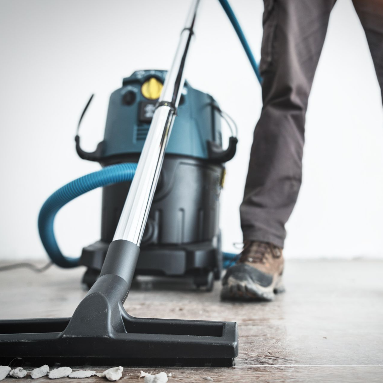 Une personne passe l'aspirateur sur un sol clair avec un aspirateur industriel bleu et noir.