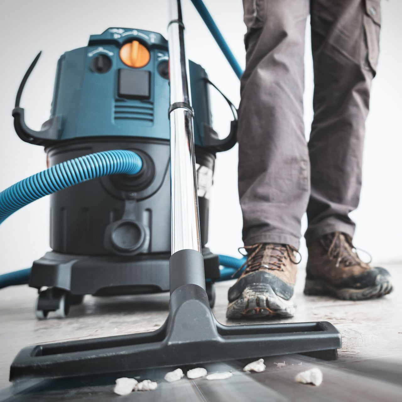 Une personne portant des bottes de travail passe l'aspirateur sur une surface à l'aide d'un gros aspirateur d'atelier bleu.