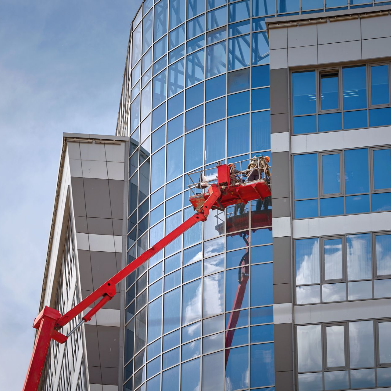 Une nacelle élévatrice rouge nettoie les vitres d'un immeuble moderne en verre ; le ciel bleu se reflète dans le ciel.