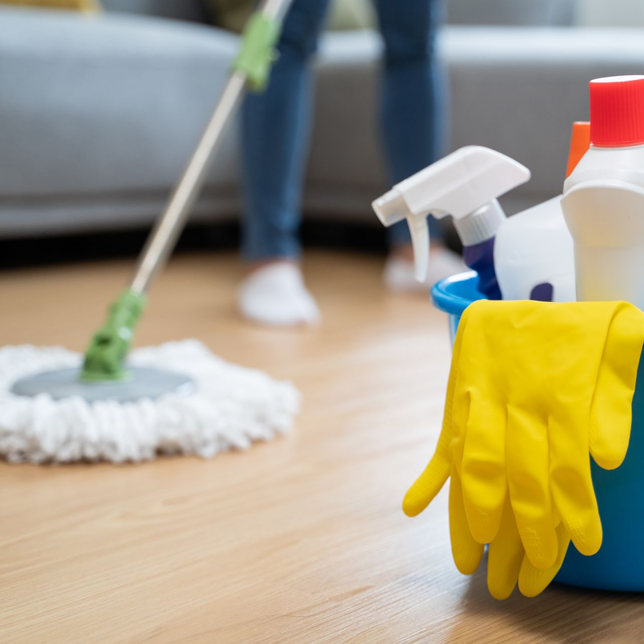 Personne passant la serpillière sur un parquet ; produits de nettoyage dans un seau bleu avec des gants jaunes.