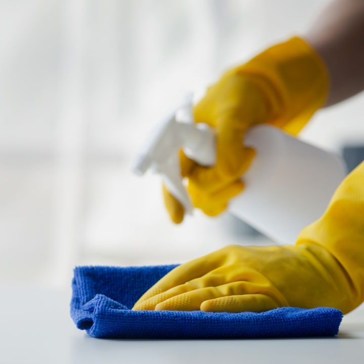 Des mains gantées de jaune vaporisent du produit nettoyant sur un chiffon bleu, nettoyant une surface blanche.