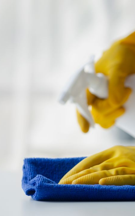 Une main gantée de jaune vaporise du nettoyant sur un chiffon en microfibre bleu posé sur une surface blanche, près d'une fenêtre.