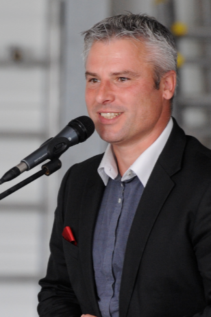 Un homme en veste de costume parle dans un microphone, souriant, avec des cheveux gris et une pochette rouge.