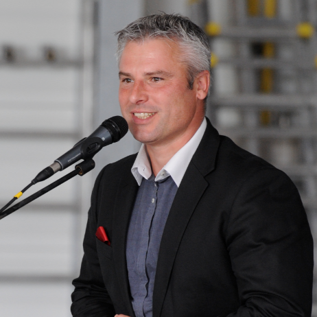 Un homme en costume parle dans un microphone, col blanc ouvert sur une chemise bleue, pochette rouge.