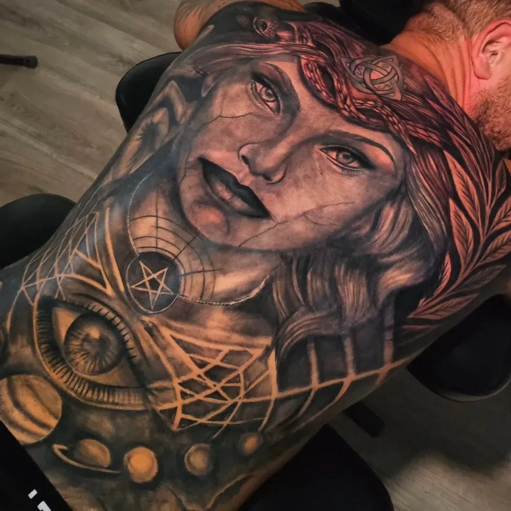 Tatuaje detallado en la espalda: rostro de mujer con patrones geométricos, un ojo y objetos celestes.