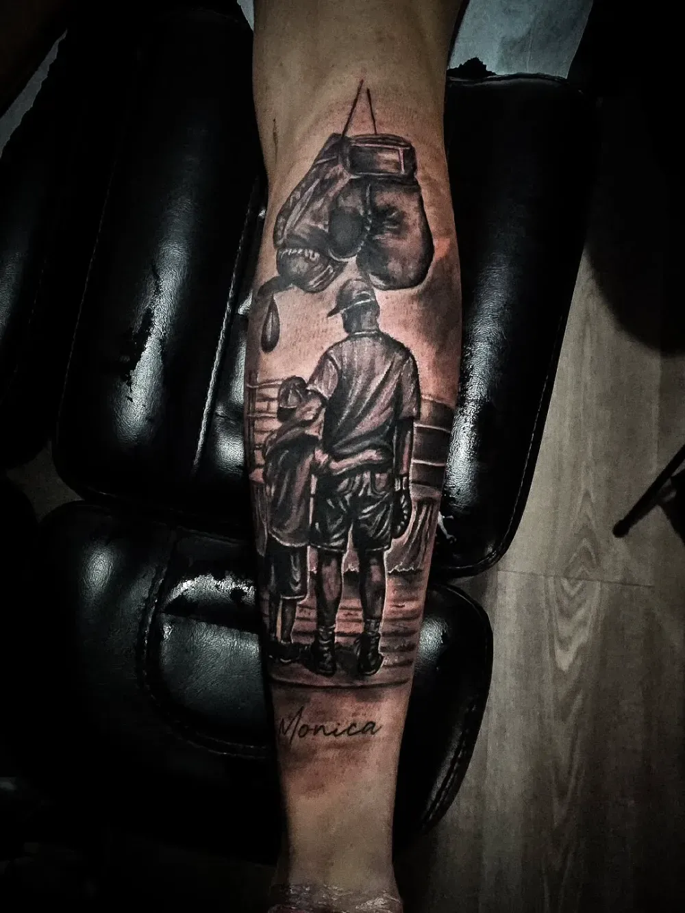 Tatuaje de una persona y un niño en un ring de boxeo, con guantes de boxeo colgando encima. "Mónica" está debajo.