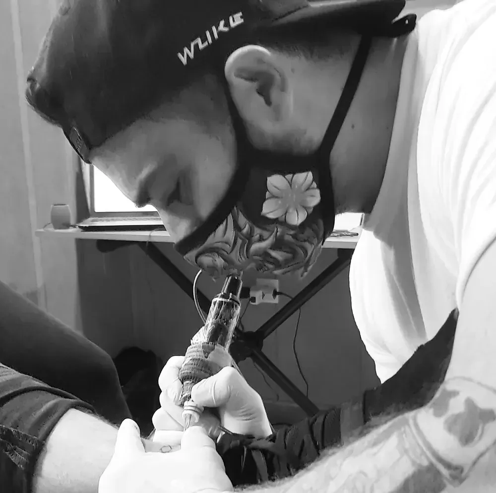 Un tatuador con máscara tatúa el tobillo de una persona. Lleva guantes y gorra.