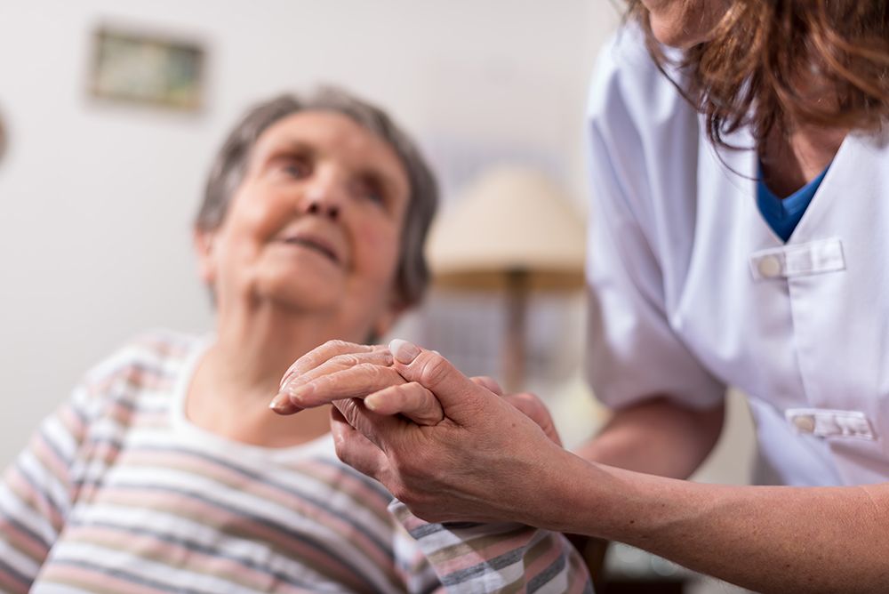 Eine Pflegekraft hält sanft die Hand einer älteren Person in häuslicher Umgebung.
