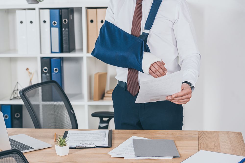 Eine Person in Geschäftskleidung mit einem Arm in einer blauen Schlinge steht an einem Schreibtisch in einem Büro und prüft Dokumente.