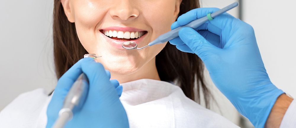 Ein Zahnarzt mit blauen Handschuhen untersucht während einer Kontrolluntersuchung die Zähne eines Patienten mithilfe eines Spiegels und zahnärztlicher Instrumente.