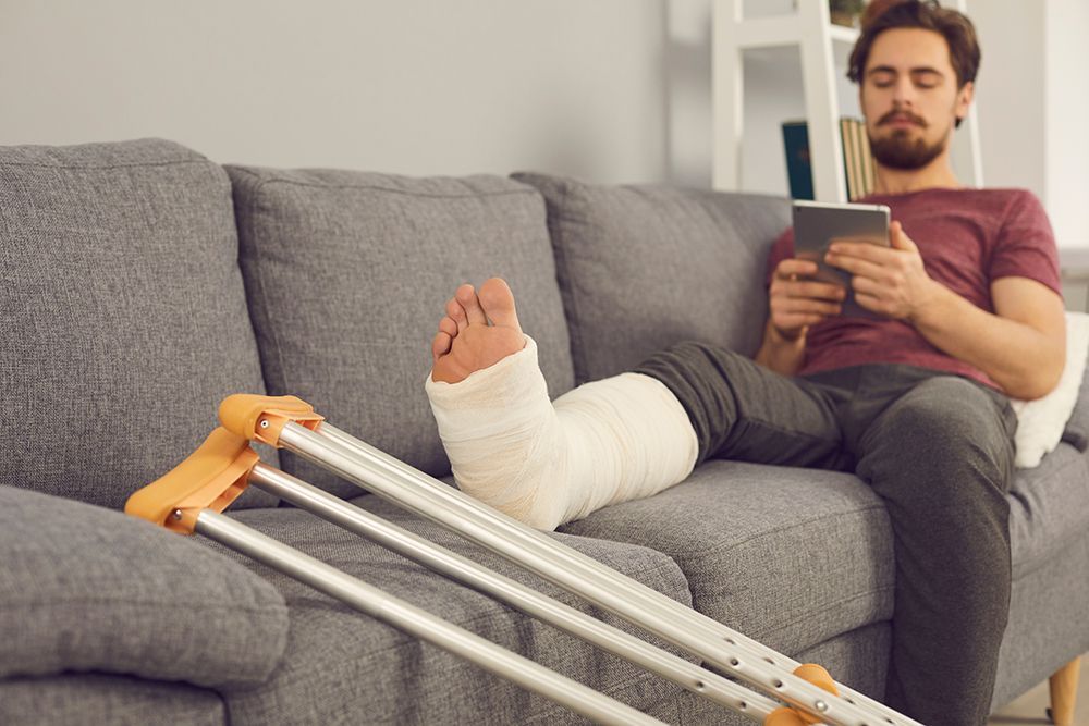 Eine Person mit einem Gipsbein ruht auf einem grauen Sofa und hält ein Tablet in der Hand, während daneben ein Paar Krücken steht.