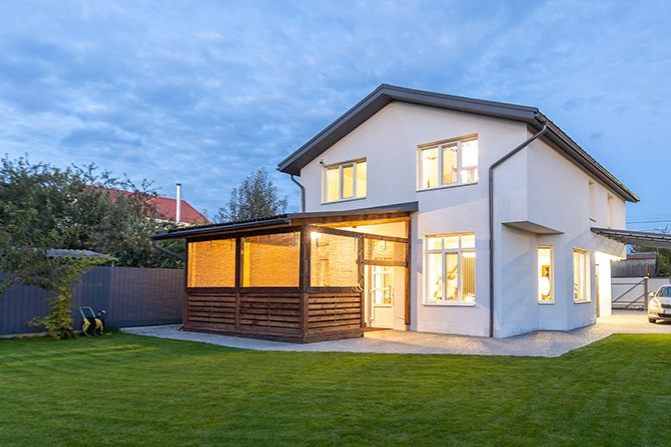 Ein modernes, weißes zweistöckiges Haus mit einer in der Abenddämmerung beleuchteten Holzveranda, vor dem Hintergrund eines grünen Rasens und blauen Himmels.