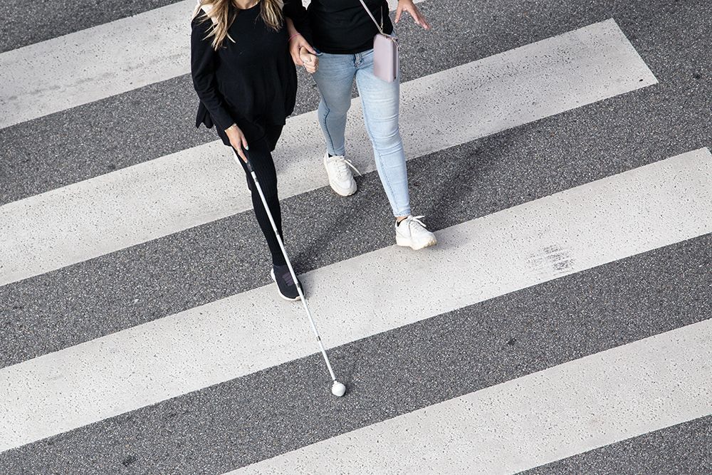 Zwei Personen überqueren einen weiß gestreiften Zebrastreifen, eine von ihnen hält einen langen weißen Gehstock.