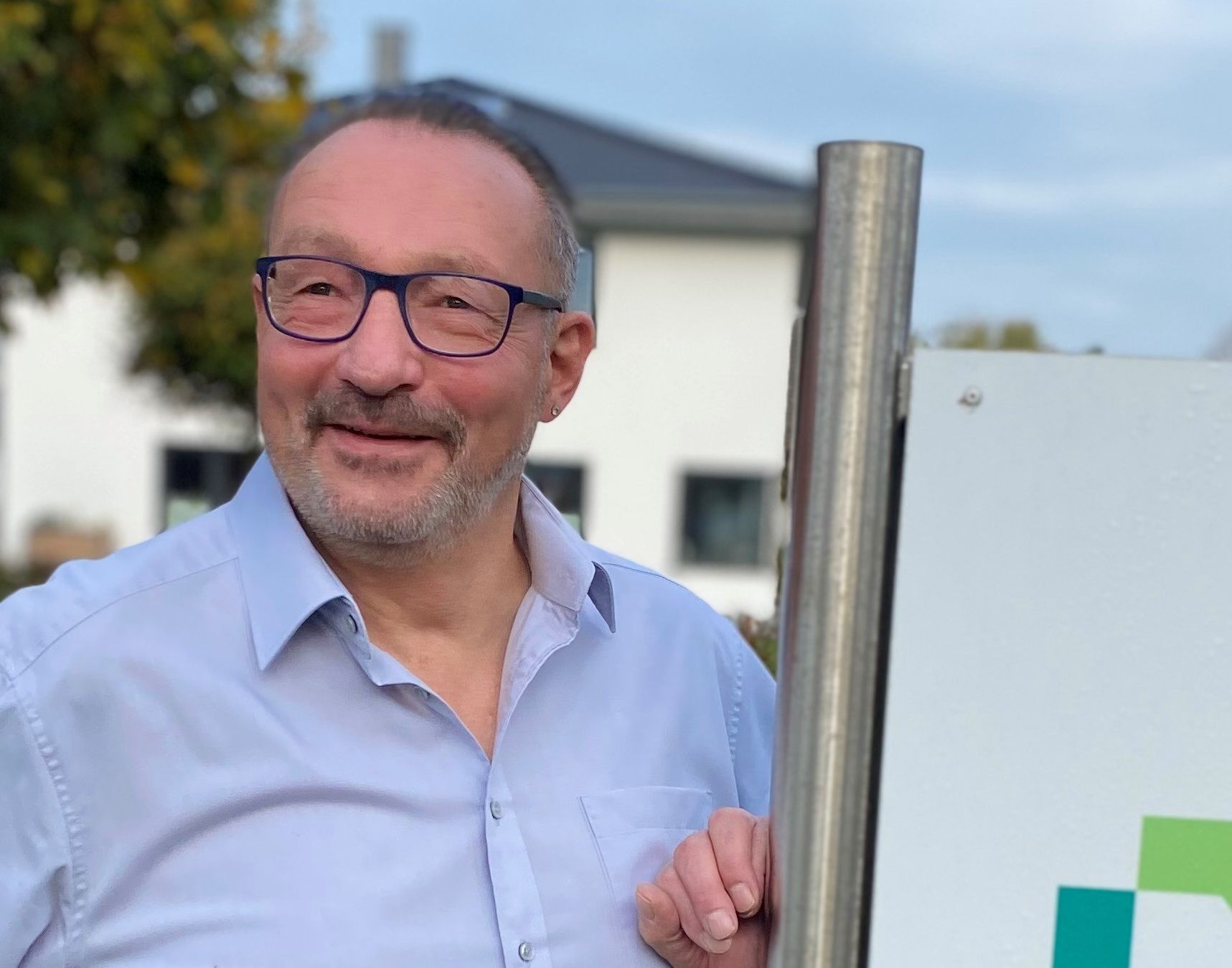 Ein Mann mit Brille und blauem Hemd steht neben einem Schild.