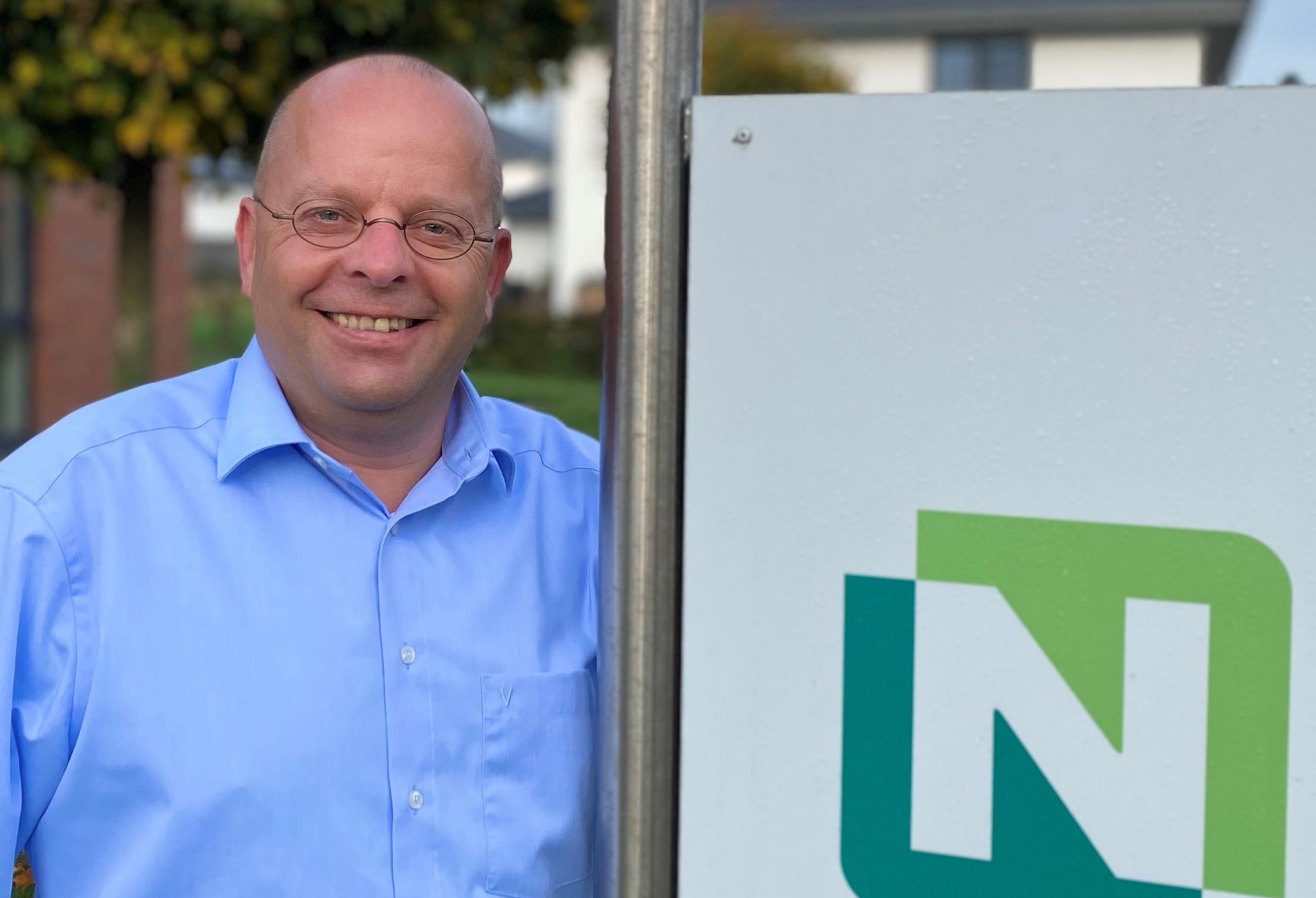 Ein Mann in blauem Hemd lächelt neben einem Schild mit grünem „N“-Logo.