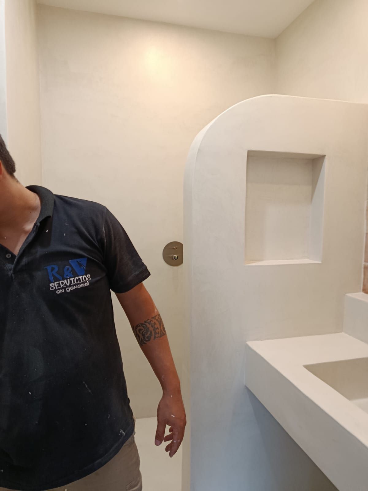 Un hombre con una camisa negra está en un baño blanco con ducha y lavabo incorporados.