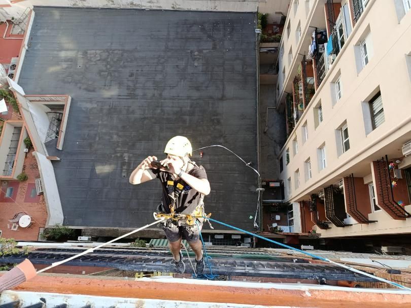 Persona descendiendo en rápel por un edificio, con casco y arnés de seguridad, entre dos edificios.