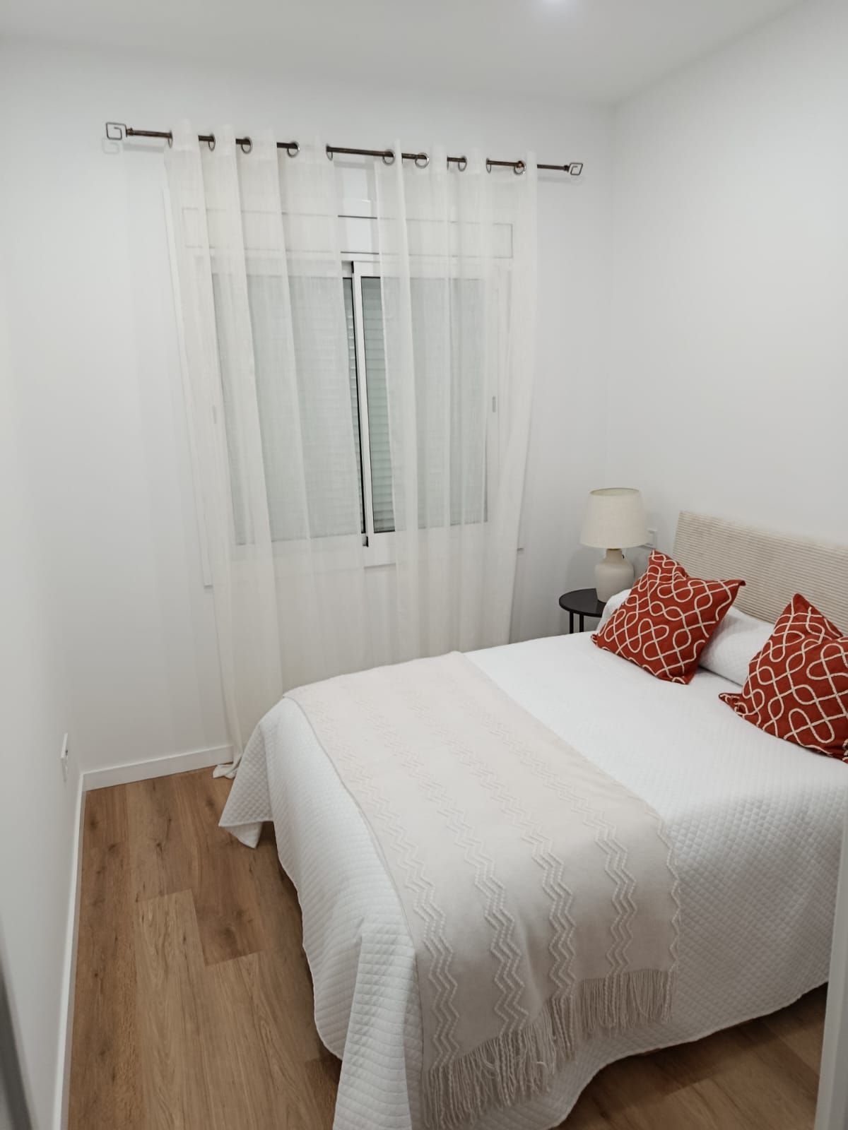 Dormitorio con paredes blancas, una cama con almohadas rojas y una ventana con cortinas transparentes.