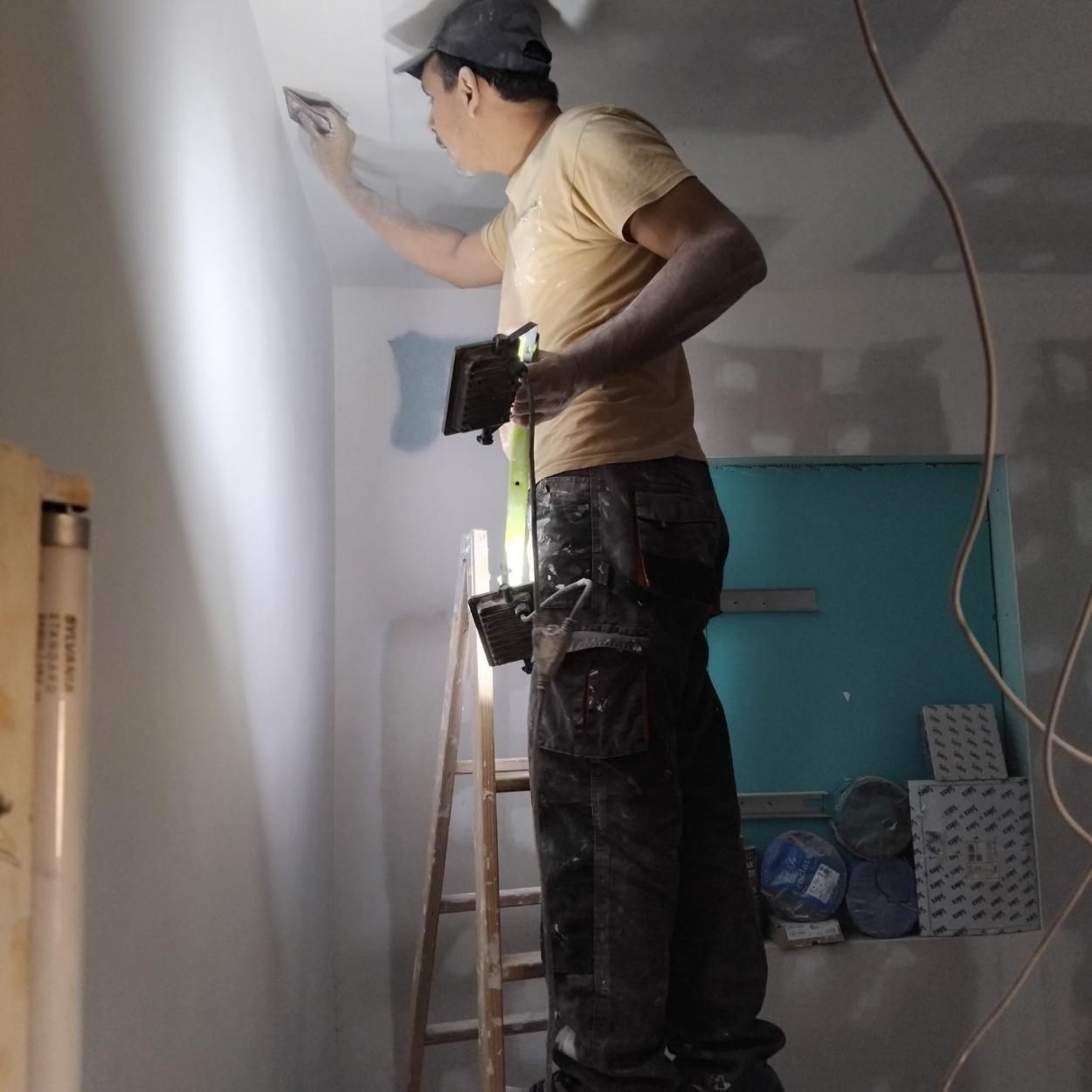 Hombre lijando una pared, de pie en una escalera en una habitación, sosteniendo una luz y una herramienta.