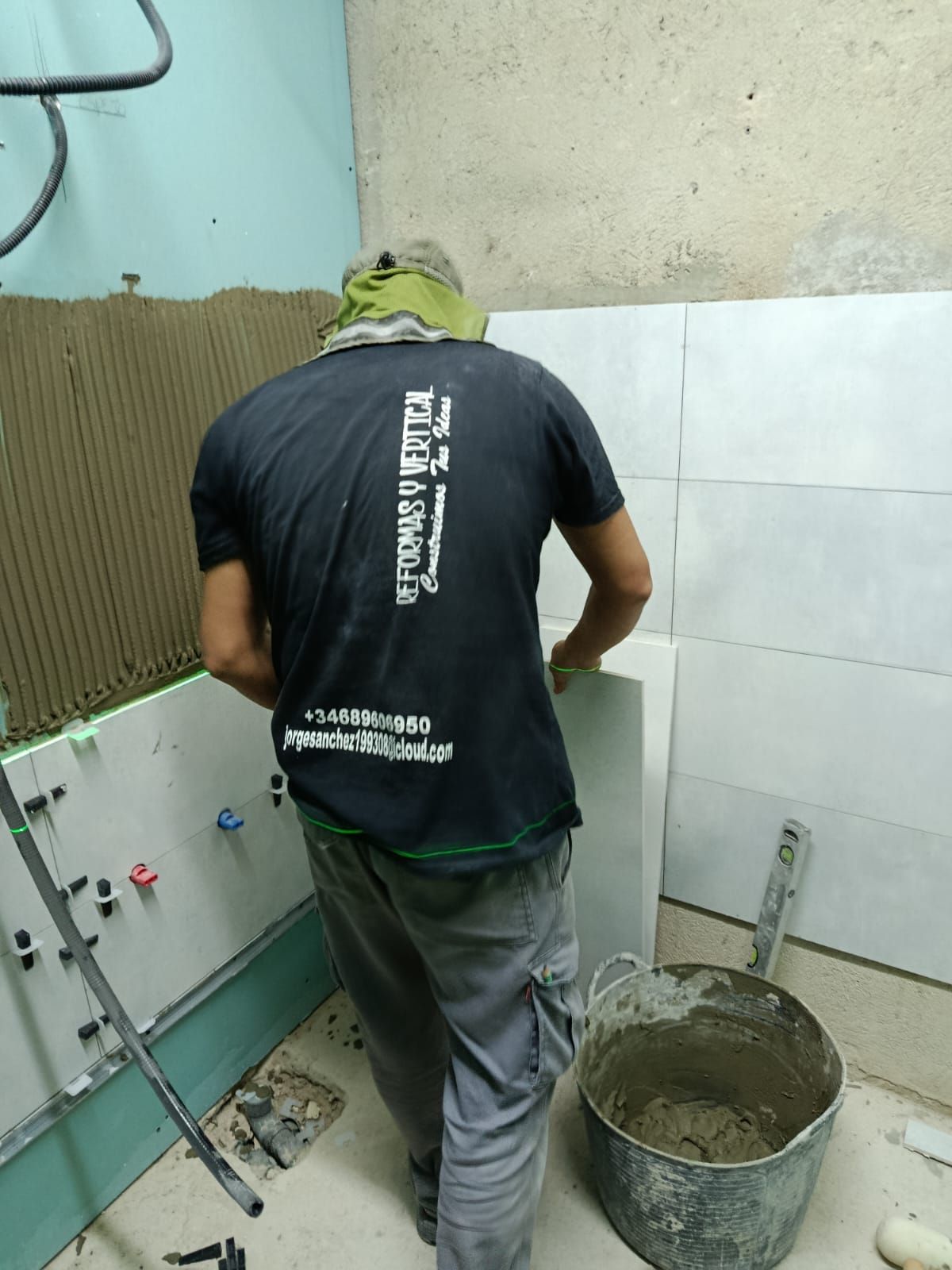 Hombre alicatando una pared en un baño. Lleva camisa negra, pantalones y sombrero. Delante hay un cubo con mortero.