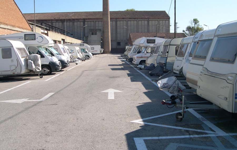 Filas de caravanas aparcadas en un terreno pavimentado, con un edificio industrial al fondo.