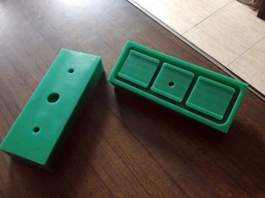Piezas de plástico rectangulares de color verde sobre una superficie de madera, una con agujeros y la otra con tres cuadrados empotrados.
