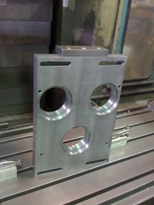 Placa de metal mecanizada con tres agujeros grandes y una pieza rectangular más pequeña sobre la mesa de una fresadora.