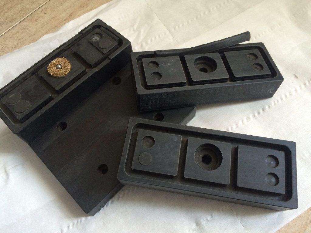 Piezas de máquina de caucho negro con áreas rebajadas y accesorios de tornillos, esparcidas sobre papel blanco.