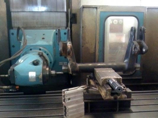 Máquina CNC con una varilla de metal sujeta en una prensa; cabezal de herramienta en el fondo, configurando un taller de máquinas.