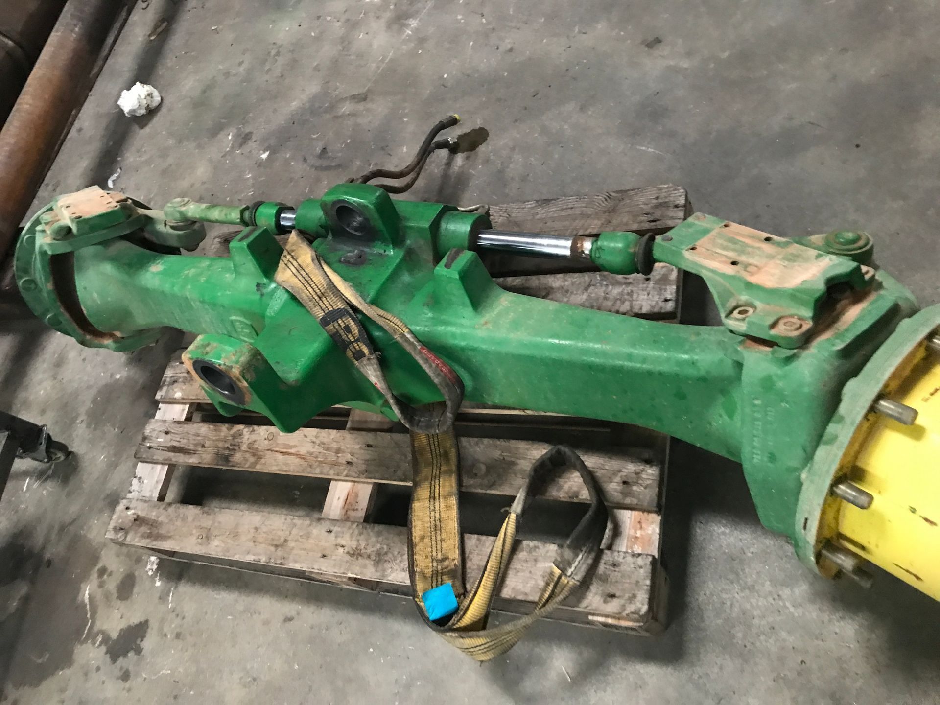 Eje de tractor John Deere verde sobre palet de madera, asegurado con una correa.